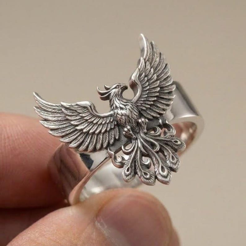 May include: A silver ring featuring a detailed, three-dimensional phoenix with wings spread wide. The metal has an oxidized finish to highlight the intricate feather textures and the bird's form, which sits atop a smooth, polished silver band.
