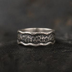 Op de afbeelding: Een zilveren ring met een getextureerde, donkergrijze band. De ring heeft een golvend ontwerp aan de boven- en onderranden, dat het getextureerde middenstuk omlijst. De ring wordt op een donkere ondergrond getoond.