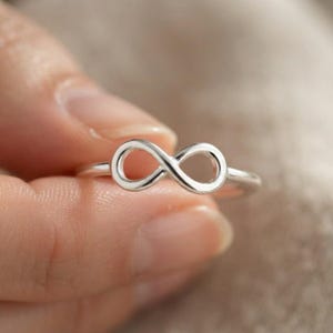 May include: A polished silver ring featuring a prominent infinity symbol design centred on the band. The metal has a smooth, reflective surface that catches the light, showcasing the continuous loop shape of the infinity motif clearly.
