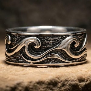Puede incluir: Un anillo de plata con un patrón de olas en relieve continuo que rodea toda la banda. El diseño incluye crestas de olas pulidas y elevadas sobre un fondo oscuro, hundido y texturizado que resalta el movimiento fluido de las formas.