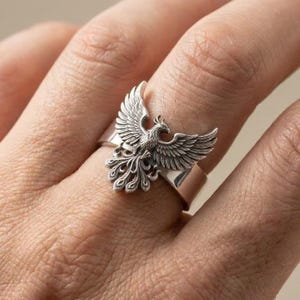 May include: A silver-toned ring featuring a detailed, raised phoenix motif with spread wings and a fanned tail. The bird design is centered on a smooth, polished band that wraps around the finger, showcasing intricate feather textures and metallic highlights.