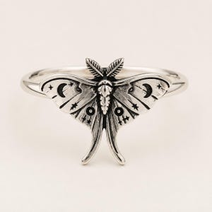 May include: A silver ring featuring a detailed moth centerpiece. The moth wings are engraved with celestial motifs including crescent moons, stars, and circular patterns. The metal has a polished finish with darkened recessed areas to highlight the intricate wing textures and the central body structure of the insect.