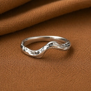 Puede incluir: Un anillo de plata de ley con un diseño de onda orgánico único. El anillo tiene una superficie texturizada y un acabado pulido, que refleja la luz. El anillo está sobre un fondo marrón. Una pieza de joyería.