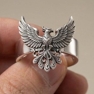 May include: A polished silver ring featuring a detailed, three-dimensional phoenix with wings spread wide and a long, ornate tail. The metal has darkened recessed areas to highlight the intricate feather textures and the bird's sharp, defined features.