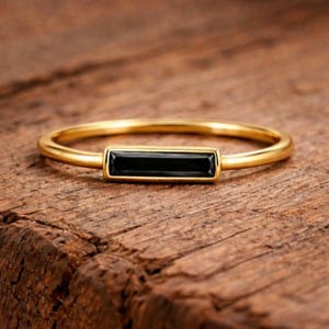 May include: A gold-colored ring with a rectangular black stone set in the center. The ring band is thin and smooth, and the stone is a deep, solid black. The ring is displayed on a wooden surface.