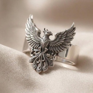 May include: A silver-toned ring featuring a detailed, three-dimensional phoenix with outstretched wings and a fanned tail. The metal has an oxidized finish in the recessed areas to highlight the intricate feather textures and the bird's form.