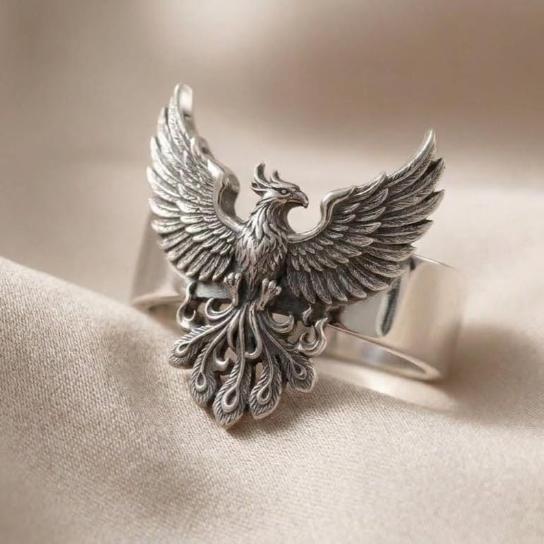May include: A silver-toned ring featuring a detailed, three-dimensional phoenix with outstretched wings and a fanned tail. The metal has an oxidized finish in the recessed areas to highlight the intricate feather textures and the bird's form.