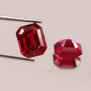 May include: Two vibrant red gemstones with an emerald cut, featuring faceted surfaces that reflect light. One gemstone is held by silver-toned metal tweezers, while the other rests nearby, showcasing deep crimson hues and a rectangular shape.