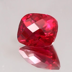 Peut inclure: Une pierre précieuse rubis taillée en coussin. La gemme est d'un rouge vif, avec de multiples facettes qui reflètent la lumière. La pierre est présentée sur une surface réfléchissante, créant une image miroir.
