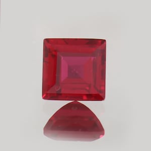 Puede incluir: Una gema roja vibrante de corte cuadrado con una superficie facetada. Debajo, una gema triangular refleja el color y la forma de la gema cuadrada. Las gemas se colocan sobre un fondo claro, resaltando su rico color y claridad.