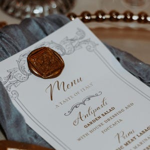 Puede incluir: Una mesa formal con una carta de menú. La carta de menú es blanca con texto dorado y un sello de cera. Una servilleta gris está debajo del menú. El plato tiene un borde dorado con cuentas y una cuchara dorada.