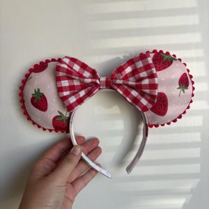 May include: Strawberry-themed mouse ears with a red and white gingham bow. The ears are pink with red strawberry designs and a red pom-pom trim. The headband is white.
