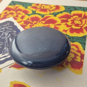 May include: A smooth, circular, dark navy blue ceramic knob with a glossy, reflective finish. The object has a slightly domed top surface and a solid, uniform color, appearing as a functional hardware piece for cabinetry or furniture drawers.