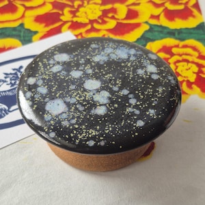 May include: A round ceramic lid with a glossy black glaze featuring speckled white and light blue spots. The underside of the lid is made of unglazed, textured brown clay. The lid has a smooth, slightly domed top surface and a flat edge.