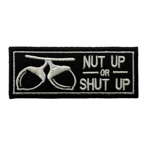 May include: A rectangular black embroidered patch featuring white stitching. On the left, there is an outline of two acorns on a branch. On the right, the text reads NUT UP OR SHUT UP in a bold, capitalized, sans-serif font style.