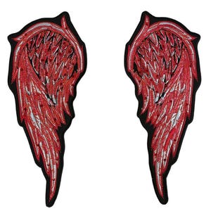 Peut inclure: Une paire d'écussons brodés en forme d'ailes d'ange. Chaque aile présente un motif de plumes détaillé avec des fils rouges et argentés, le tout entouré d'une bordure noire solide qui souligne la forme complète des ailes.