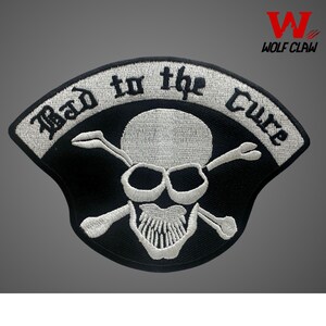 May include: A black and white embroidered patch featuring a skull and crossbones design. The text "Bad to the Cure" is stitched in a curved white banner above the skull. The patch has a black border and a black background behind the skull.
