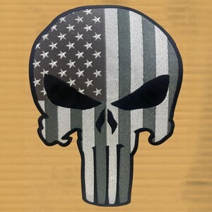 May include: An embroidered patch in the shape of a skull with a stylized American flag design. The left side features white stars on a dark gray field, while the right side and lower portion consist of alternating vertical gray and white stripes.