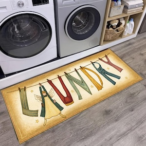 May include: A rectangular floor mat with a beige background featuring the word LAUNDRY spelled out with clothing items hanging on a clothesline. An angel figure forms the letter L, followed by red pants, green pants, yellow fabric, and blue overalls.
