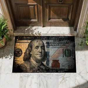 Può includere: Uno zerbino rettangolare con una stampa grafica di una banconota da cento dollari americani. Il design mostra il ritratto di Benjamin Franklin, il testo "The United States of America" e "One Hundred Dollars" in uno stile usurato e invecchiato.