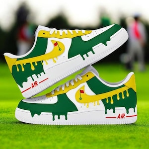 May include: A pair of white trainers with green and yellow golf-themed design. The trainers feature a white sole, green and yellow paint drip design, and a golf flag logo. The word "AIR" is printed in red on the side of the trainers.