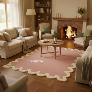 May include: A living room scene featuring a pink rug with a scalloped cream border. The rug sits on a hardwood floor, surrounded by beige sofas, armchairs, and a wooden coffee table. A fireplace with a burning fire and a bookshelf are in the background.