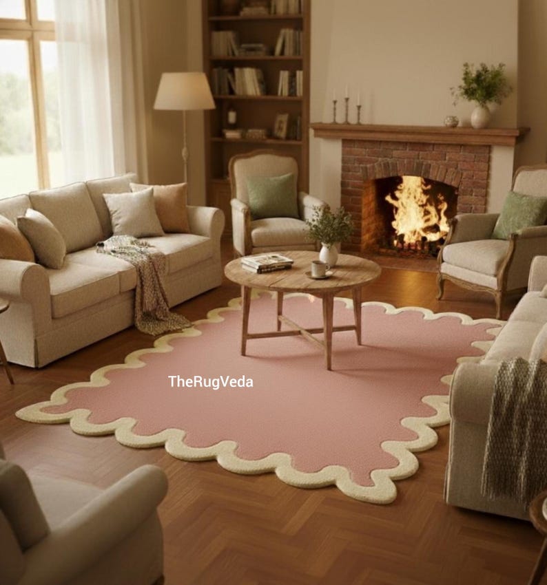 May include: A living room scene featuring a pink rug with a scalloped cream border. The rug sits on a hardwood floor, surrounded by beige sofas, armchairs, and a wooden coffee table. A fireplace with a burning fire and a bookshelf are in the background.