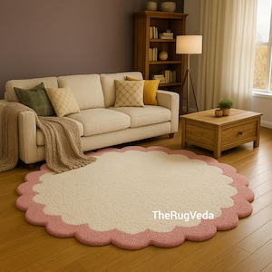 May include: A round, cream-colored rug with a scalloped pink border. The rug is placed on a hardwood floor in a living room setting, with a beige sofa, throw pillows, and a wooden coffee table. The rug's design adds a soft, decorative element to the space.
