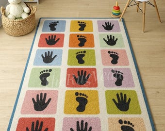 May include: A rectangular children's rug with a white background and a blue border. The rug features colorful squares with black handprints and footprints. The rug is placed on a light wood floor.