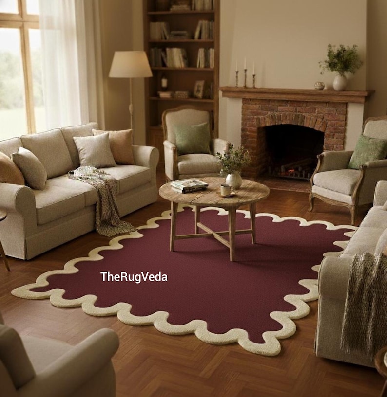 May include: A burgundy area rug with a scalloped, cream-colored border. The rug is in a living room setting with beige sofas, chairs, a wooden coffee table, and a fireplace. The text "TheRugVeda" is visible on the rug.