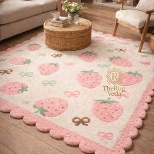 May include: A cream-colored area rug featuring a repeating pattern of pink strawberries and small bows in shades of green and brown. The rug has a scalloped pink border and includes the text "R TheRug Veda" printed in gold on the surface.