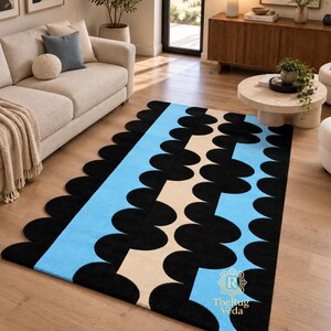 May include: A rectangular area rug featuring a scalloped edge design with vertical stripes in black, light blue, and beige. The rug displays a repeating pattern of black semi-circles along the borders and center, with the text TheRug Veda in gold.