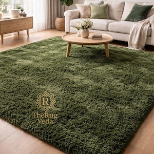 May include: A rectangular, deep olive green shag rug with a thick, plush pile texture. The rug features a gold-colored logo emblem printed on the surface with the text TheRug Veda, displayed in a decorative frame with an ornate R symbol.