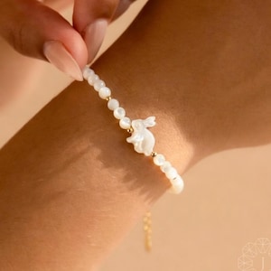 Può includere: Un delicato braccialetto con un ciondolo a forma di coniglietto bianco e piccole perline bianche rotonde. Il braccialetto ha dettagli color oro e una delicata catena. Il braccialetto è tenuto da una persona.