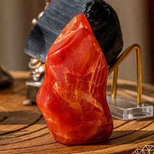 May include: A polished, flame-shaped carnelian gemstone, displaying a vibrant red-orange hue with internal veining. The stone sits on a wooden surface, with other decorative objects in the background. The gemstone is a natural, decorative item.