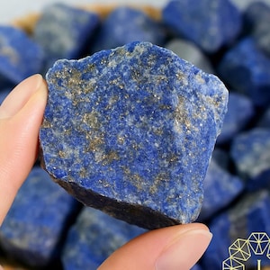 May include: A hand holds a chunk of deep blue lapis lazuli with gold flecks. Other lapis lazuli stones are in a woven basket in the background. The stone is rough and textured, with a rich color.