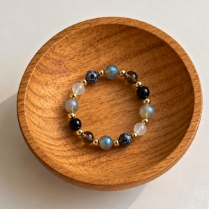 Pode incluir: Um anel circular de contas com contas redondas alternadas em tons de azul translúcido, preto opaco, branco e castanho escuro, intercaladas com pequenas contas espaçadoras metálicas em tom dourado num padrão repetitivo.