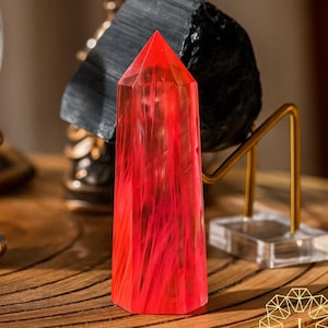 May include: A faceted, red crystal obelisk stands on a wooden surface. The crystal has a pointed top and a rectangular base. Behind it is a black, rough-textured stone and a gold-colored metal stand. The obelisk is the focal point.