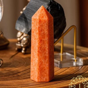 Pode incluir: Um obelisco de cristal laranja com uma superfície texturizada está sobre uma mesa de madeira. O obelisco é o ponto focal, com uma pedra cinza escuro e um suporte de metal dourado no fundo. O obelisco é um item decorativo.