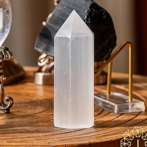 Könnte beinhalten: Ein hoher, weißer Selenit-Kristallturm mit spitzer Spitze. Der Kristall wird auf einer Holzoberfläche präsentiert, mit einem schwarzen Stein und einem goldfarbenen Metallständer im Hintergrund. Der Kristall ist etwa 15 cm hoch.