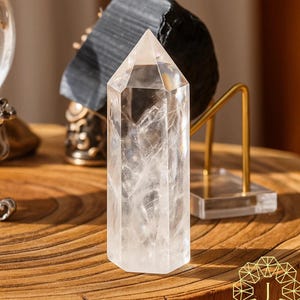 May include: A clear quartz crystal point stands on a wooden surface. The crystal is hexagonal and tapers to a point. Other decorative objects are in the background, including a black stone and a gold-coloured metal stand.