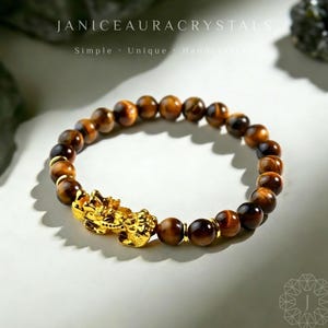 May include: A beaded bracelet featuring tiger's eye stones and a gold-colored charm. The bracelet has a mix of brown and gold beads, with a detailed gold charm. The text "JANICEAURACRYSTALS" is at the top.