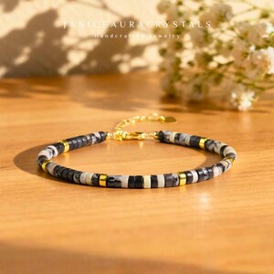 May include: A beaded bracelet with black, white, and gold accents. The bracelet features small, flat beads in a pattern of black, white, and gold. The clasp and connecting chain are gold. The text "JANICEAURACRYSTALS Handcrafted Jewelry" is visible.