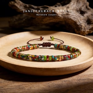 Op de afbeelding: Een armband met kleine, platte, ronde regenboogjaspis kralen in tinten rood, groen, blauw en oranje, geaccentueerd met goudkleurige metalen tussenstukjes. De armband heeft een bruin koord met een verstelbare schuifknoopsluiting.