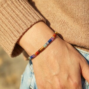 May include: A beaded bracelet featuring small, cylindrical beads in a variety of colors including purple, blue, red, brown, orange, yellow, green, and grey, arranged in a sequence along a thin, flexible cord worn around the wrist.
