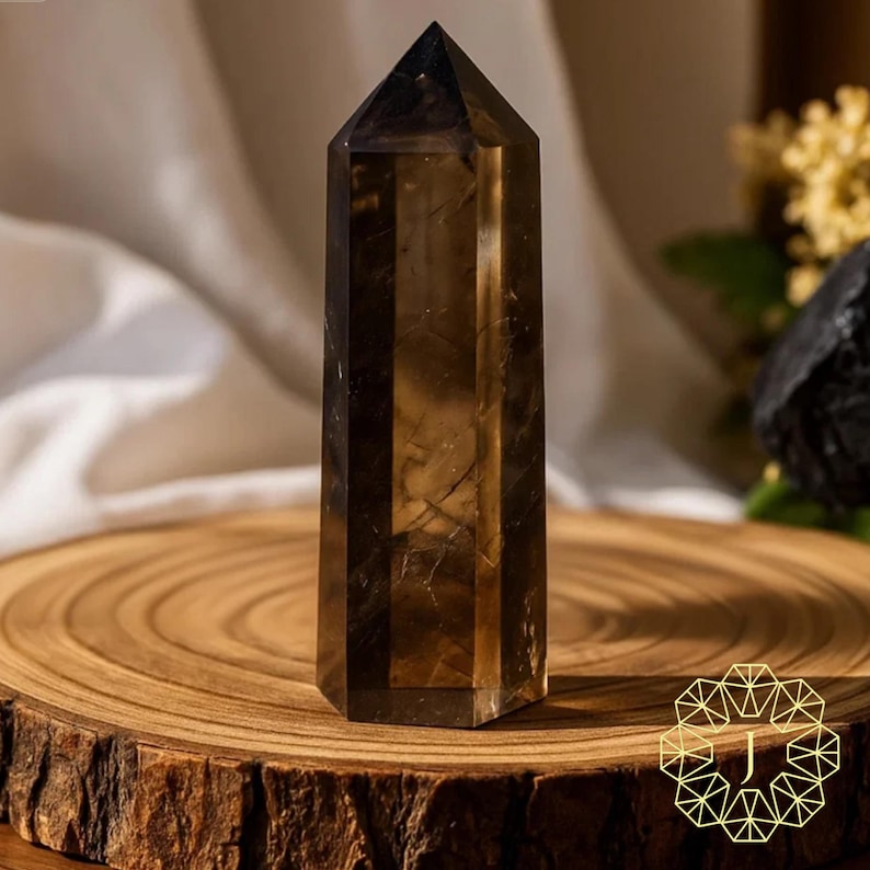 May include: A faceted, dark brown crystal point stands on a wooden slice. The crystal has a pointed top and a rectangular shape. The wood slice has visible growth rings. The background includes a white cloth and greenery.