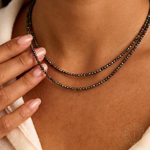 May include: Two strands of beaded necklaces. The necklaces are composed of small, dark green and burgundy beads. The necklaces are worn around the neck. The image is a close-up shot of the necklaces.