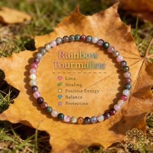 May include: A rainbow tourmaline beaded bracelet with various colors, including pink, green, and brown, is displayed on a large, dried leaf. The text "Rainbow Tourmaline" is above a list of benefits: Love, Healing, Positive Energy, Balance, and Protection.