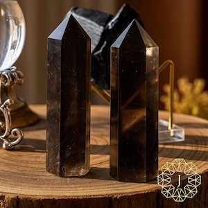 May include: Two tall, polished smoky quartz crystal points stand on a wooden surface. The crystals are dark brown with a faceted, pointed top. A clear glass sphere and a black stone are visible in the background.