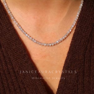 May include: A delicate necklace composed of small, round, light-colored beads. The necklace is displayed against a brown background. The text "JANICEAURACRYSTALS" and "Minimalist Jewelry" are visible.
